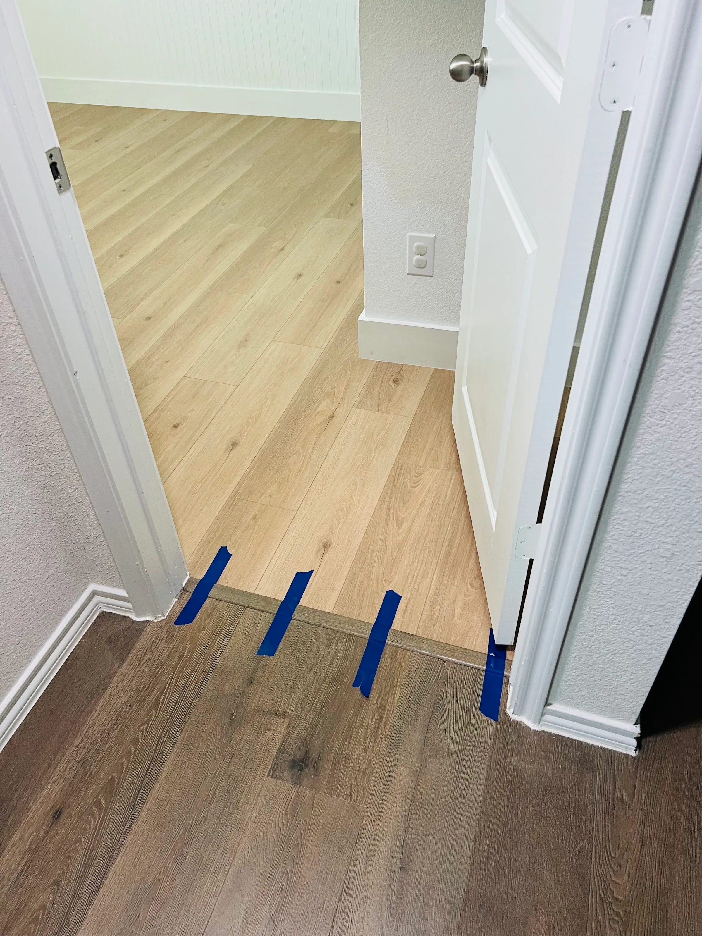 Vista de una puerta mostrando una transición de piso LVP claro dentro de una recámara al piso más oscuro del pasillo, con cinta azul a lo largo de la transición.