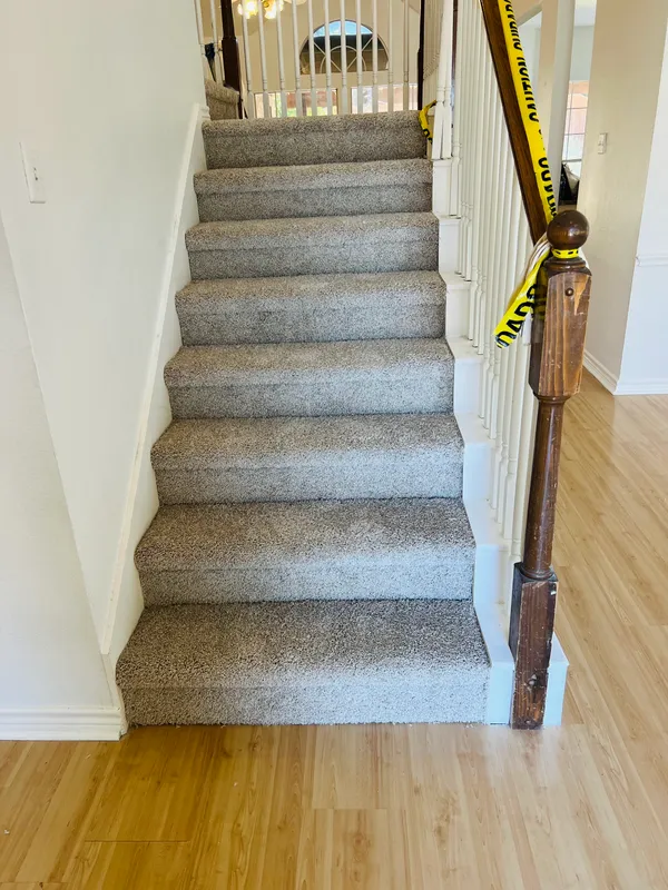 The carpeted staircase from the first floor: light gray speckled carpet on every step, a wood newel post with caution tape, and a light wood-look floor at the bottom.