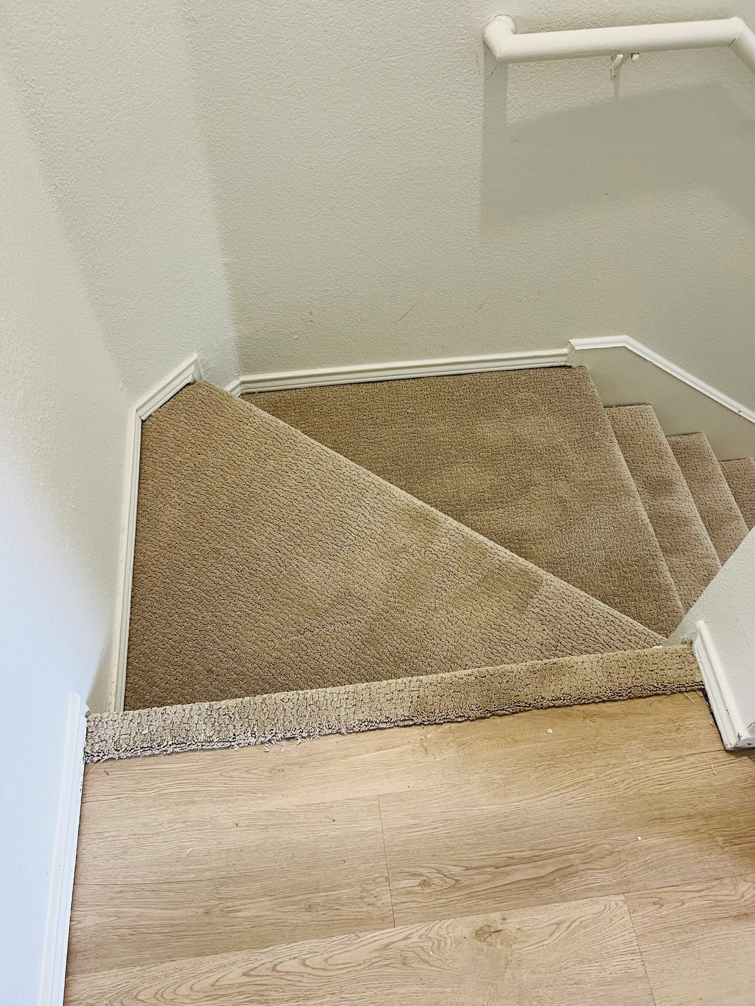 Vista hacia abajo desde el último escalón: alfombra beige sobre los escalones compensados de la vuelta, con la nariz superior terminando sobre el nuevo piso LVP claro.