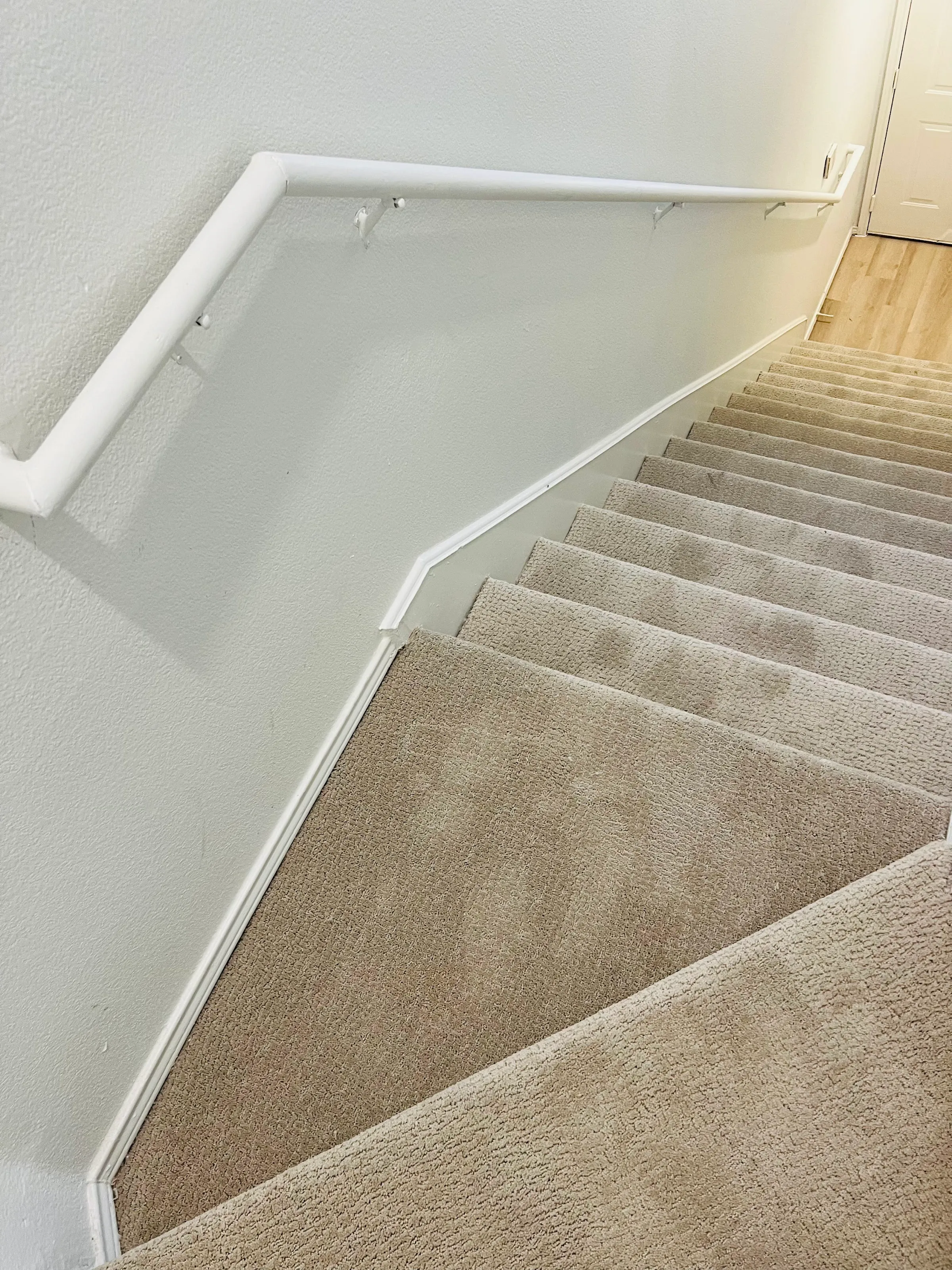 El tramo completo de escaleras alfombradas en beige visto desde arriba, con pasamanos blanco en la pared y el piso LVP claro abajo.